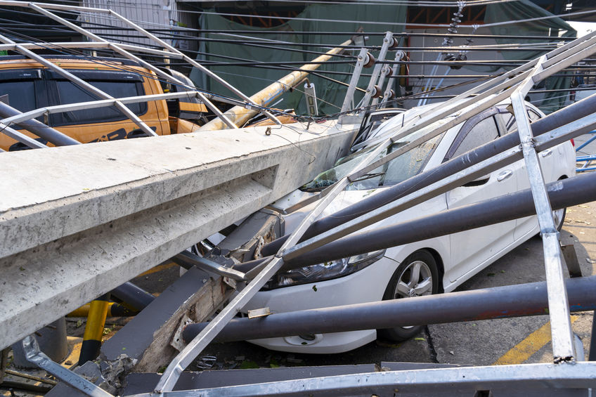 A white car is crushed under fallen power lines and poles, with a yellow vehicle in the background. Debris surrounds the area, highlighting the critical role Wind & Storm Damage Insurance Adjusters play in such disasters.