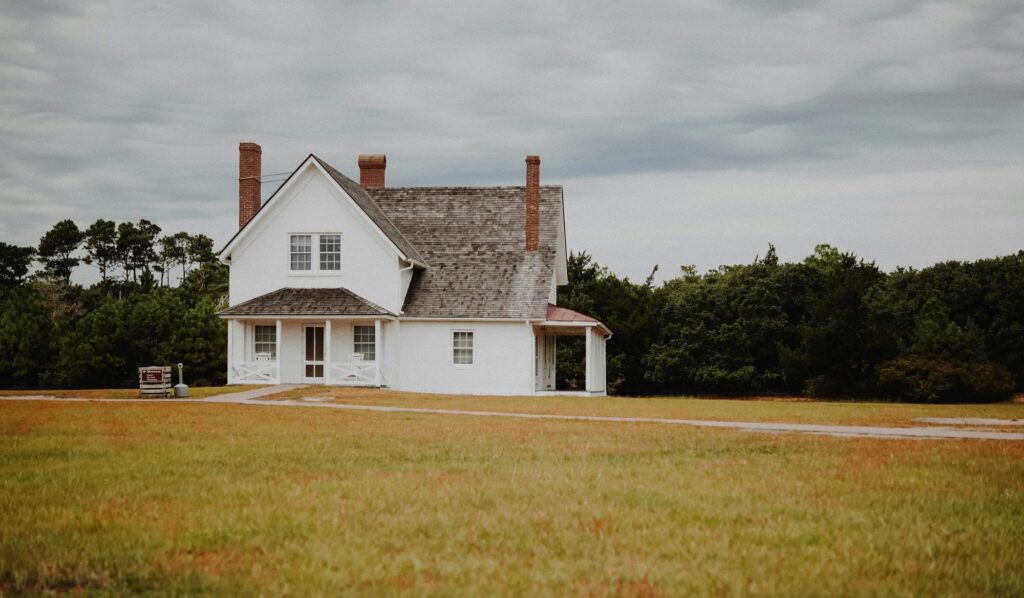 A white two-story house with multiple chimneys stands in a grassy field surrounded by trees under a cloudy sky.