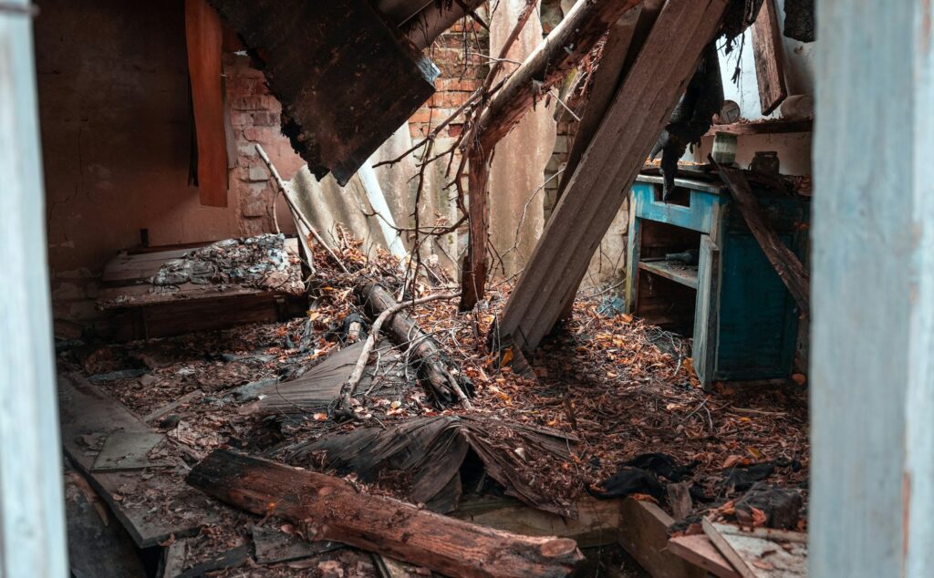 Interior of a dilapidated room with collapsed wooden beams, fallen debris, and damaged furniture. The space appears neglected and in severe disrepair.